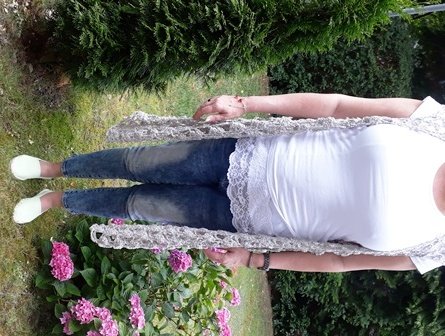 Person hält lange beigefarbene gehäkelte Weste mit Lochmuster vor Garten mit Hortensien