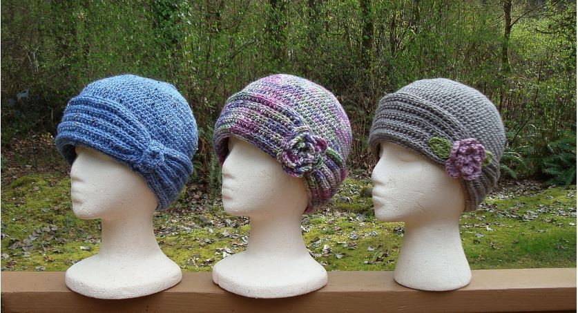 Three crochet cloche hats on mannequin heads: blue with knotted band (left), variegated purple-pink-green with flower (center), and grey with purple flower and green leaf (right).