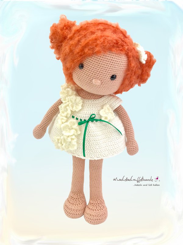 Gehäkelte Amigurumi-Puppe mit rotem lockigem Haar, cremeweißem Kleid mit Blumenapplikationen und grünem Band vor hellem Hintergrund