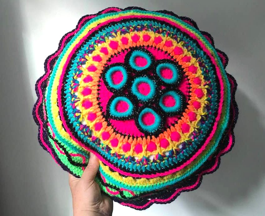 Hand hält rundes, gehäkeltes Kissen oder Kissenbezug mit buntem Mandala-Muster in leuchtenden Farben.