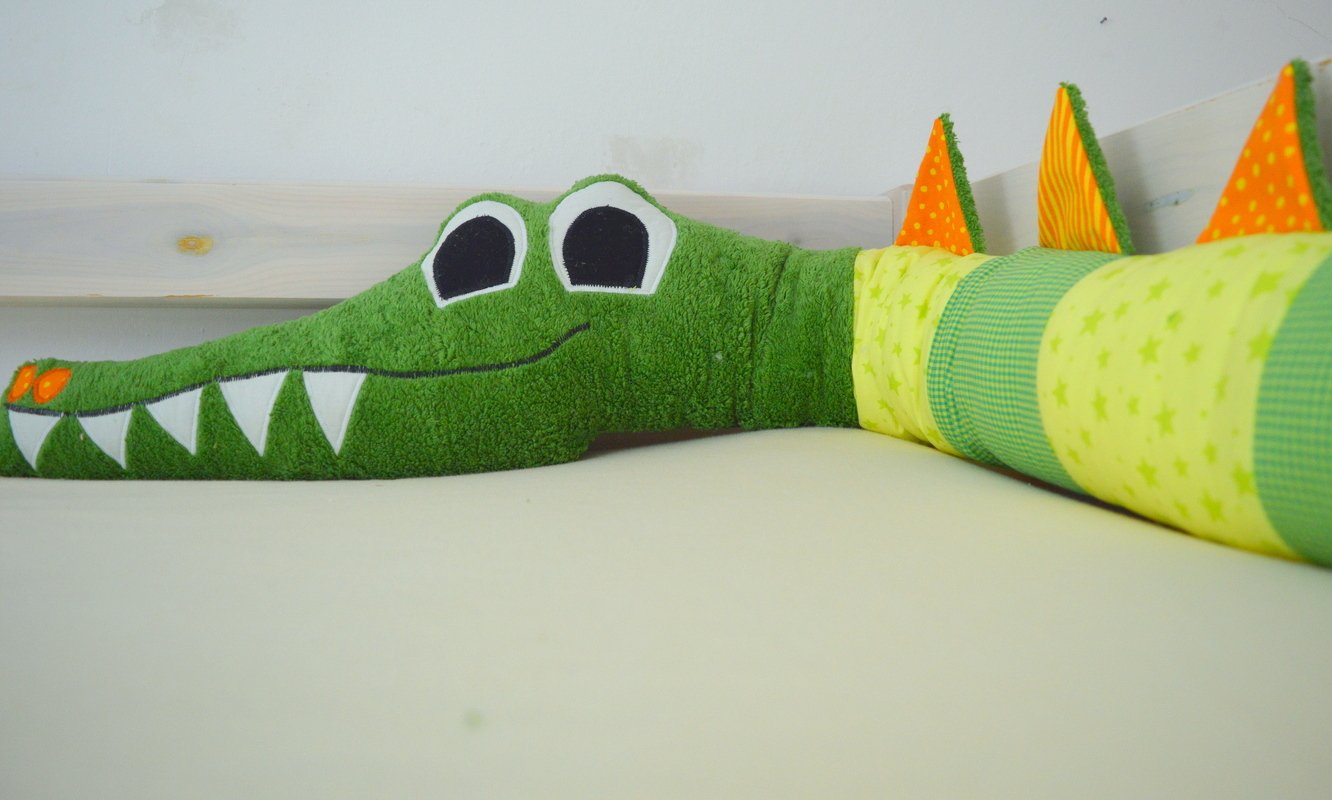 Langes grünes Stoffkrokodil mit großen Augen, weißen Zähnen und bunten Zacken auf hellem Untergrund.