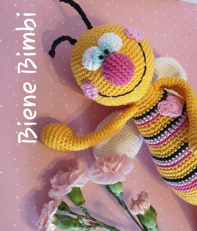 Gehäkelte Amigurumi-Biene gelb-schwarz-pink mit weißen Flügeln auf rosa gepunktetem Hintergrund neben rosa Nelken