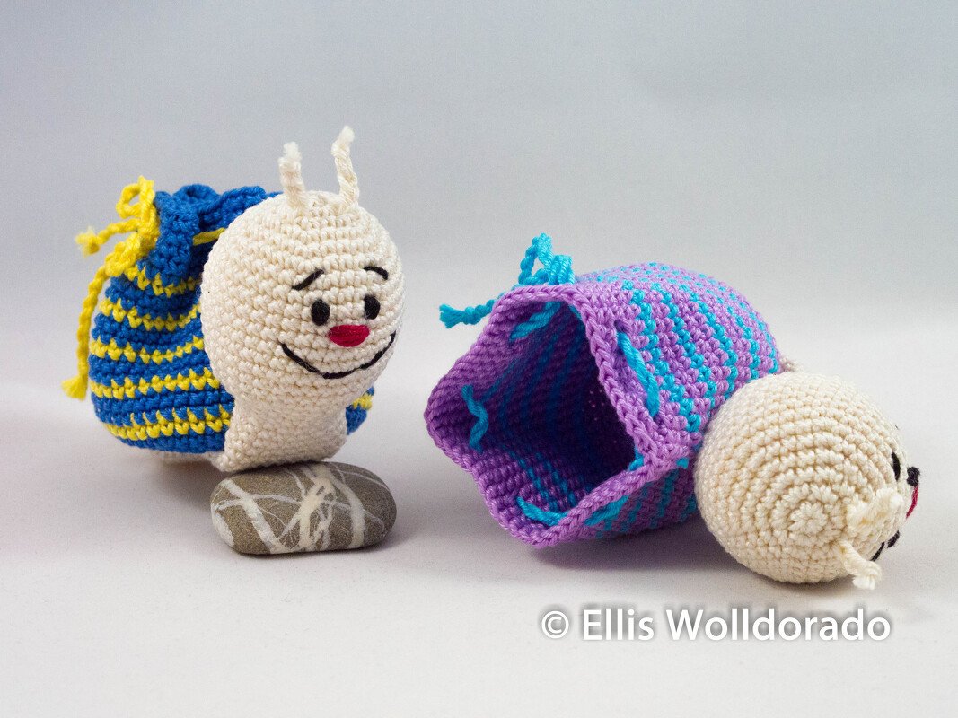 Zwei gehäkelte Amigurumi-Schnecken mit gestreiften Beutel-Schneckenhäusern; eine steht auf einem Stein, die andere liegt offen daneben.