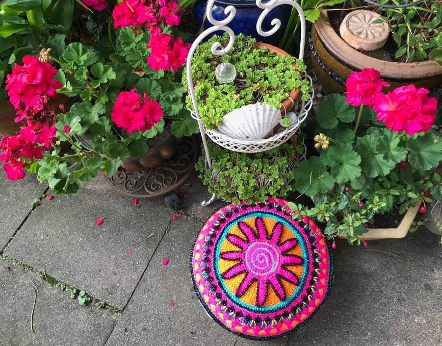 Rundes gehäkeltes Kissen mit Sonnenmotiv in Pink, Orange und Türkis auf grauen Pflastersteinen vor pinken Geranien