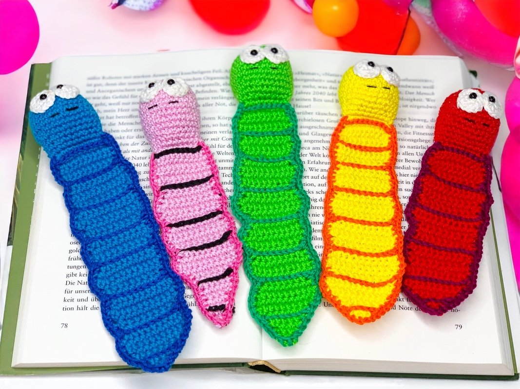 Five crocheted worm bookmarks in blue, pink, green, yellow and red on an open book.