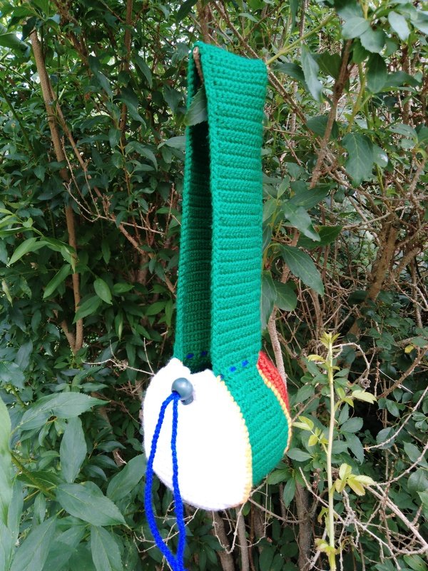 Runder Häkelbeutel mit langem Träger und blauem Kordelzug, an einem Strauch hängend