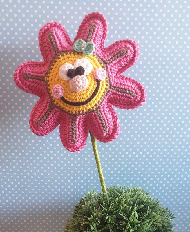 Nahaufnahme einer gehäkelten Blume mit gelbem Smiley-Mittelteil, rosa Blütenblättern, kleiner grüner Schleife und grünem Stängel vor hellblauem, gepunktetem Hintergrund.