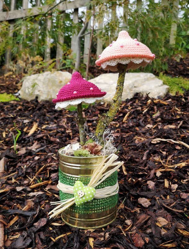 Zwei gehäkelte Pilze in Rosa und Pink auf Ästen in einer verzierten Metalldose auf Rindenmulch