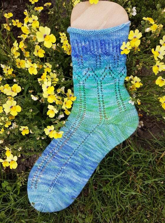 Blau-grün gestrickte Damensocke mit Ajourmuster auf einem Holzsockenformer zwischen gelben Blumen