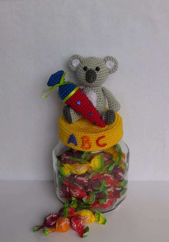 Handgehäkelter Koala mit bunter Schultüte auf gelbem gehäkeltem Deckel mit ABC-Buchstaben auf einem Glas mit Bonbons