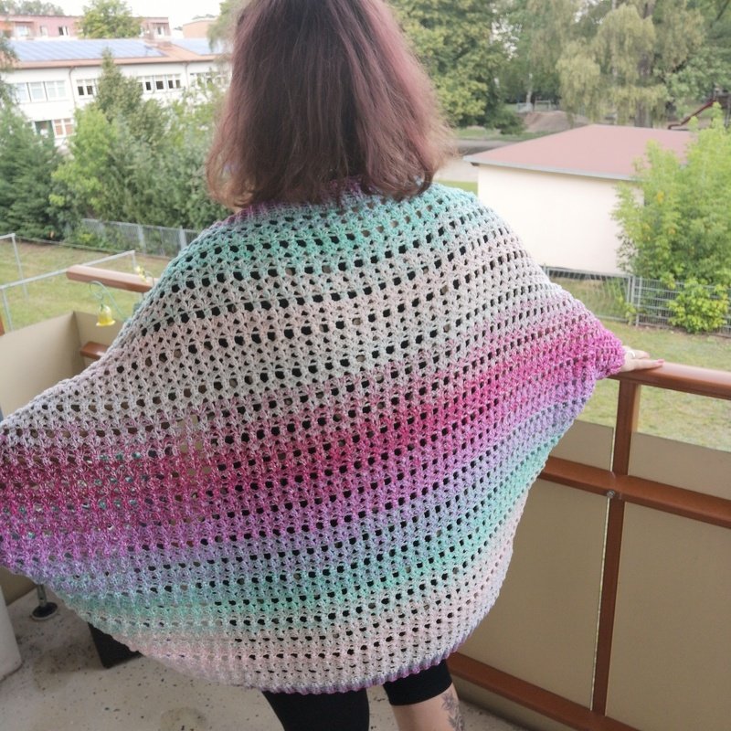 Rückenansicht einer Person mit gehäkeltem, pastellfarbenem Seelenwärmer im offenen Netzmuster auf einem Balkon