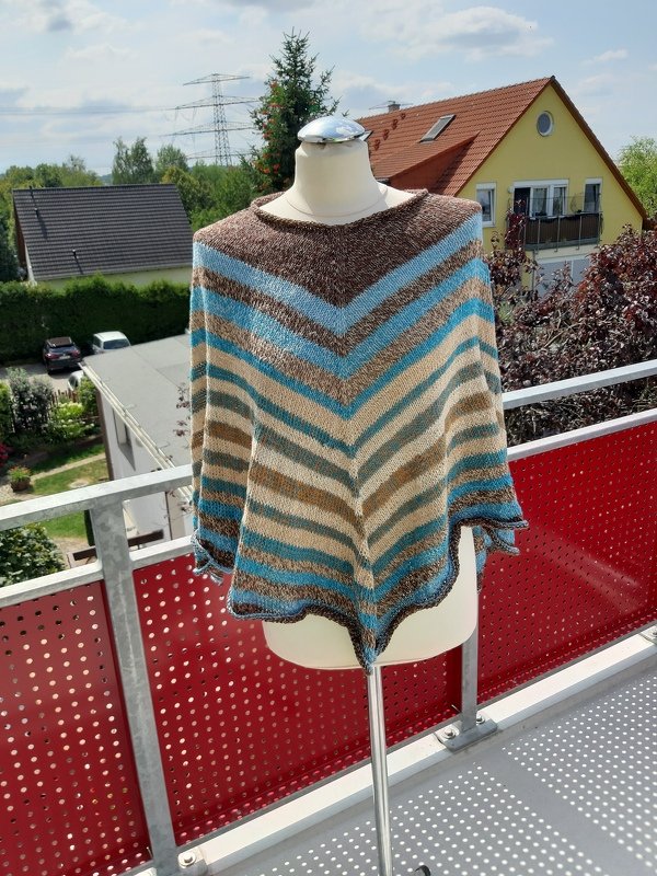 Gestrickter Damenponcho mit V‑Streifen in Braun, Beige und Blau auf einer Schneiderpuppe auf einem Balkon