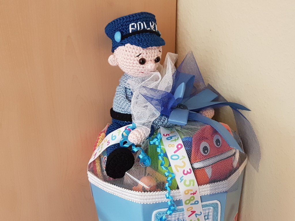Gehäkelte Amigurumi-Polizistenpuppe in blauer Uniform auf einer verzierten hellblauen Geschenkbox mit Bändern und Netzstoff.
