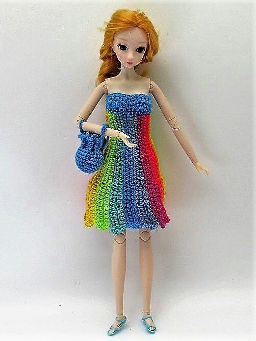 Puppe mit langem blondem Haar in gehäkeltem trägerlosem Regenbogenkleid, blauer Tasche und blauen Sandalen.