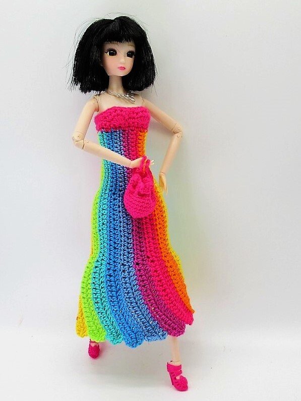 Modepuppe mit kurzem schwarzem Haar trägt ein langes, gehäkeltes Regenbogenkleid und eine kleine gehäkelte Tasche