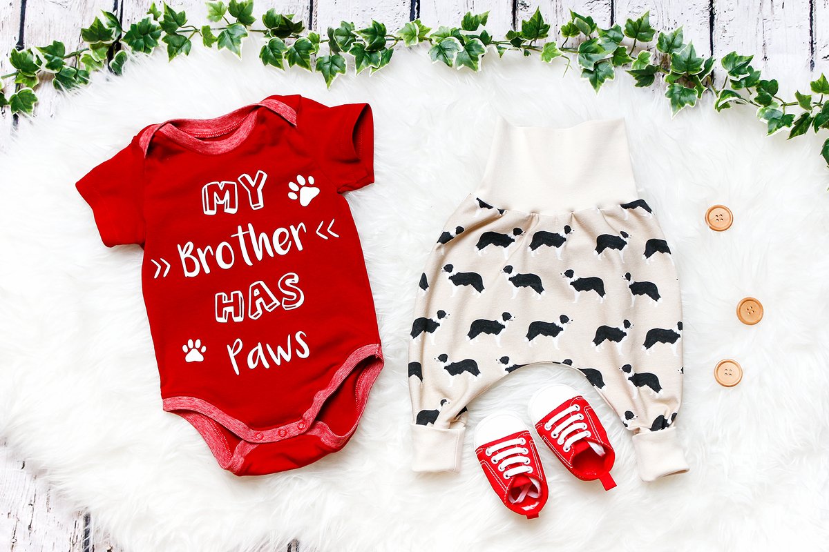 Roter Babybody mit weißem Plottmotiv 'My Brother Has Paws' neben beige Babyhose mit schwarzen Tier‑Silhouetten und roten Babyschuhen auf weißem Fell