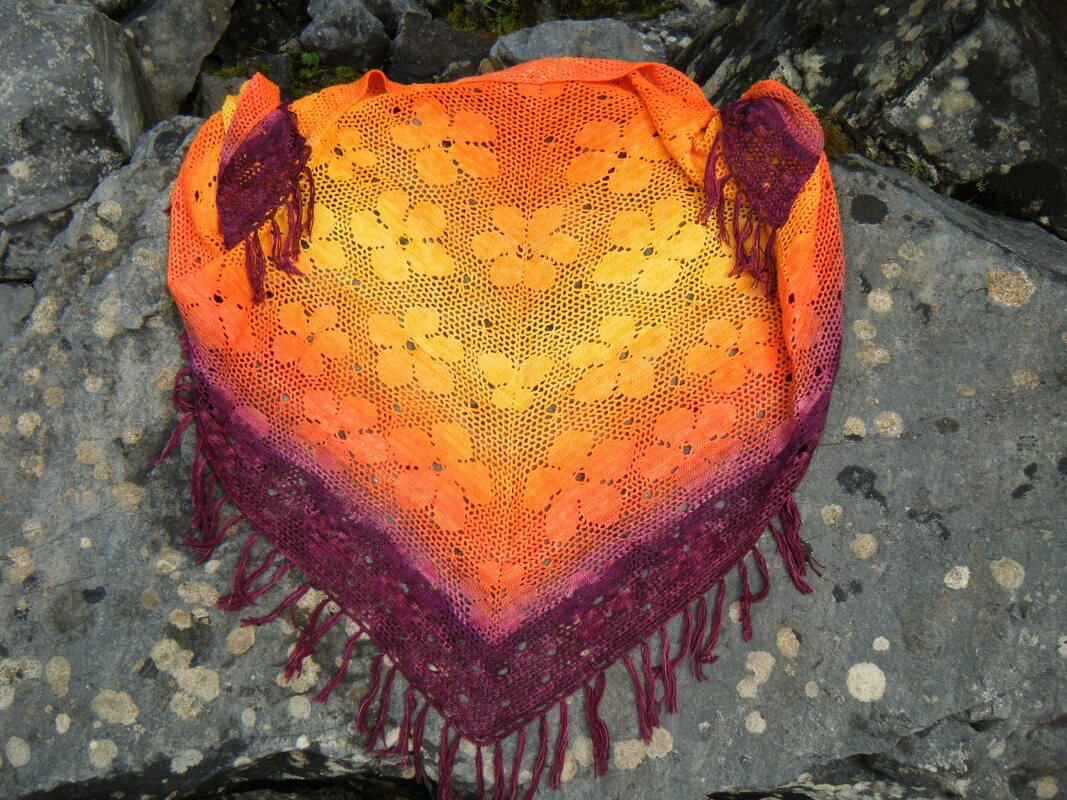 Gehäkeltes Dreieckstuch in Gelb, Orange und Violett mit Blumen-Lochmuster und Fransen auf grauem Felsen