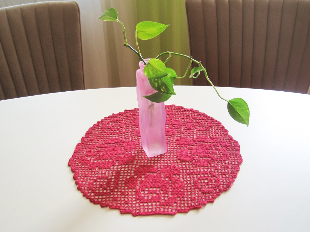 Rundes pinkes gehäkeltes Deckchen mit Rosenmuster auf weißem Tisch und rosa Vase