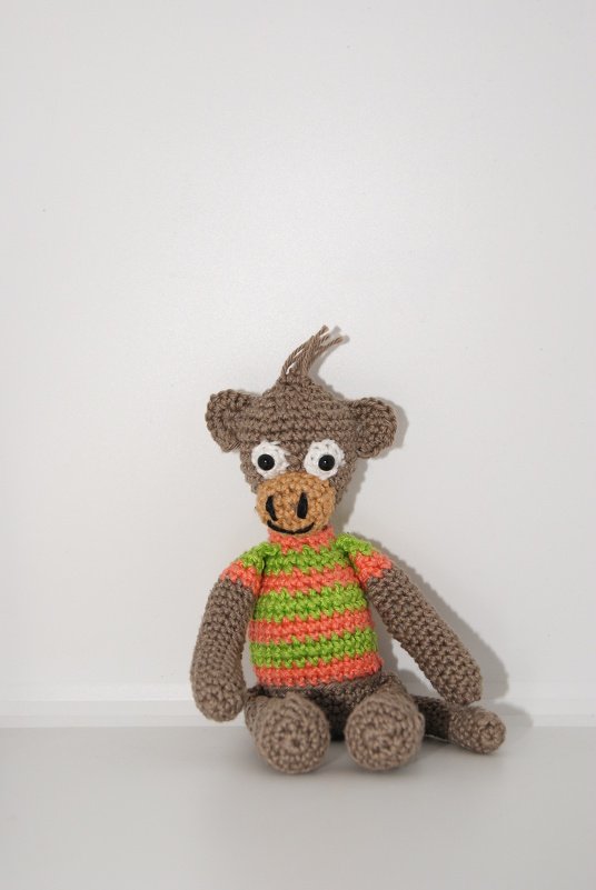 Gehäkelter brauner Amigurumi-Affe mit grün-orange gestreiftem Shirt, sitzend vor weißem Hintergrund.