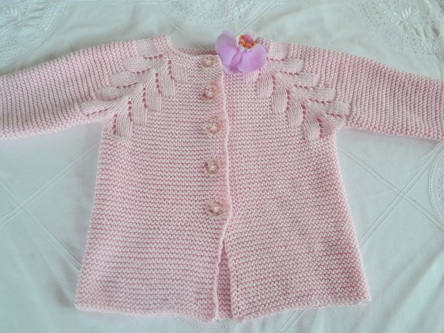 Rosa gestrickte Babyjacke mit langen Ärmeln, Knopfleiste und Blättermuster an den Schultern. Kleine lila Stoffblüte am Kragen.