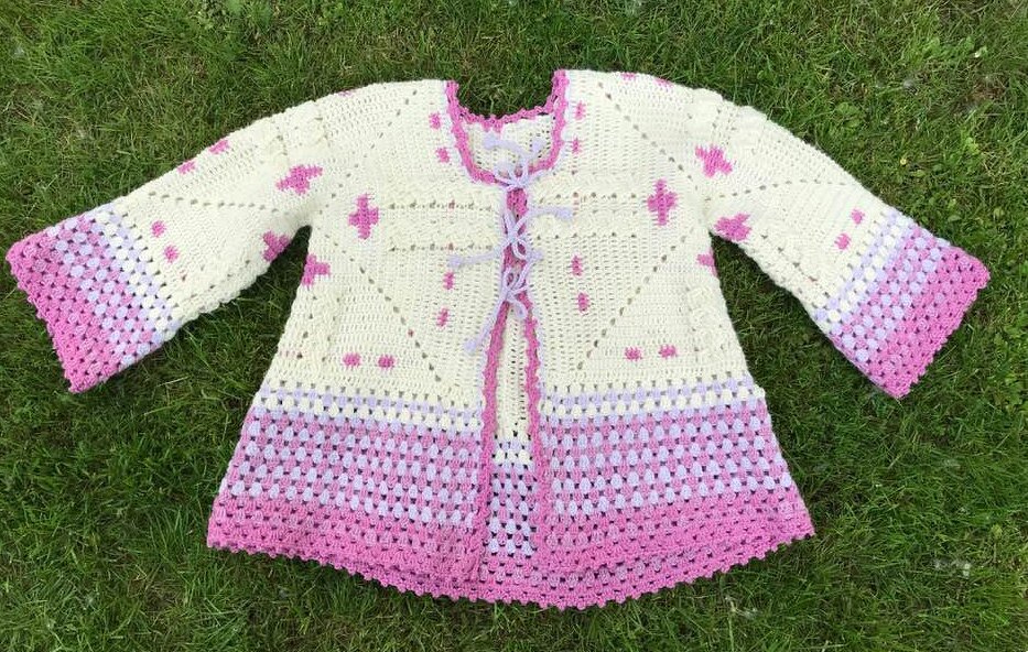Gehäkelte Damenjacke cremeweiß mit Pink-Lila Farbverlauf an Ärmeln und Saum, offener Schnitt, auf Gras.