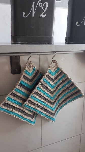 Zwei gehäkelte Topflappen mit Zickzackstreifen in Beige, Grau und Türkis hängen an Haken.