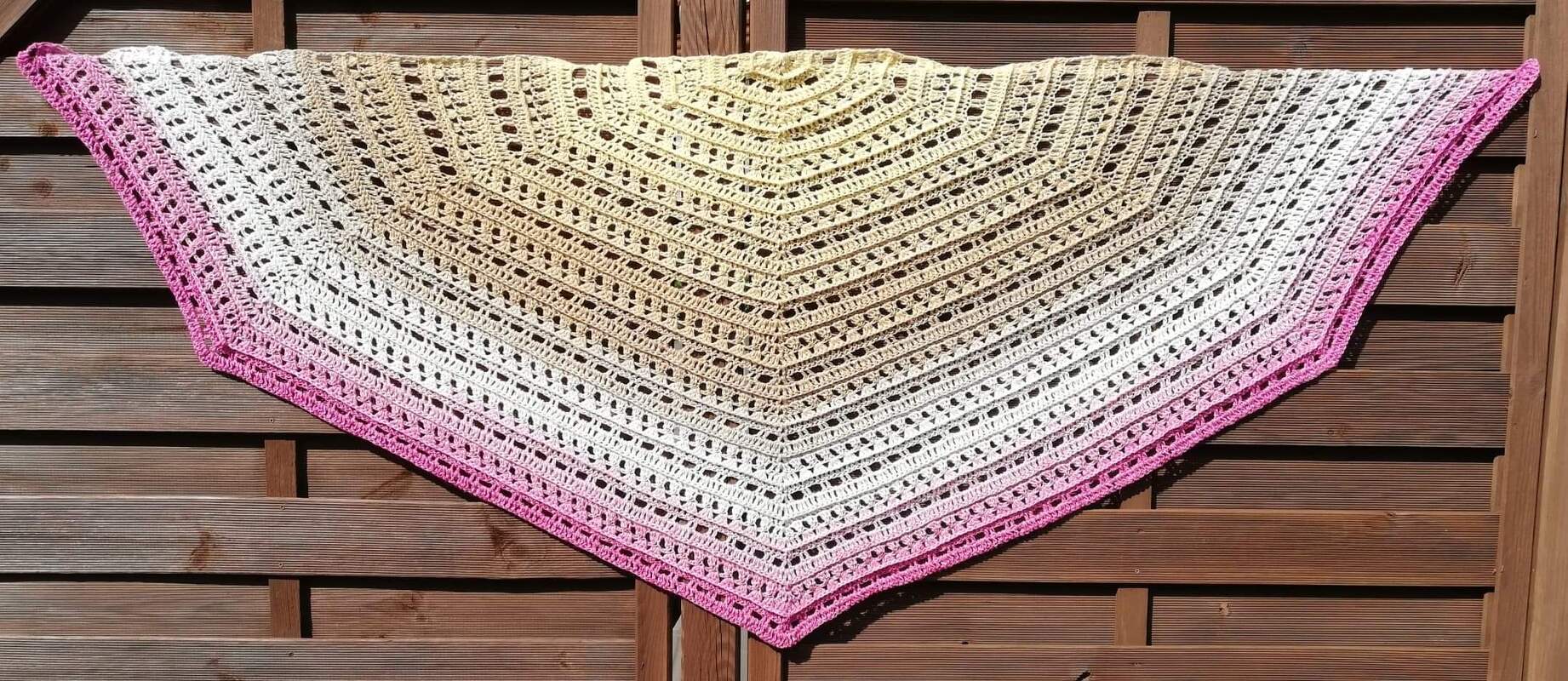 Gehäkeltes halbrundes Tuch mit Farbverlauf von Gelb zu Rosa, Lochmuster und pinker Kante, aufgehängt vor Holzlatten