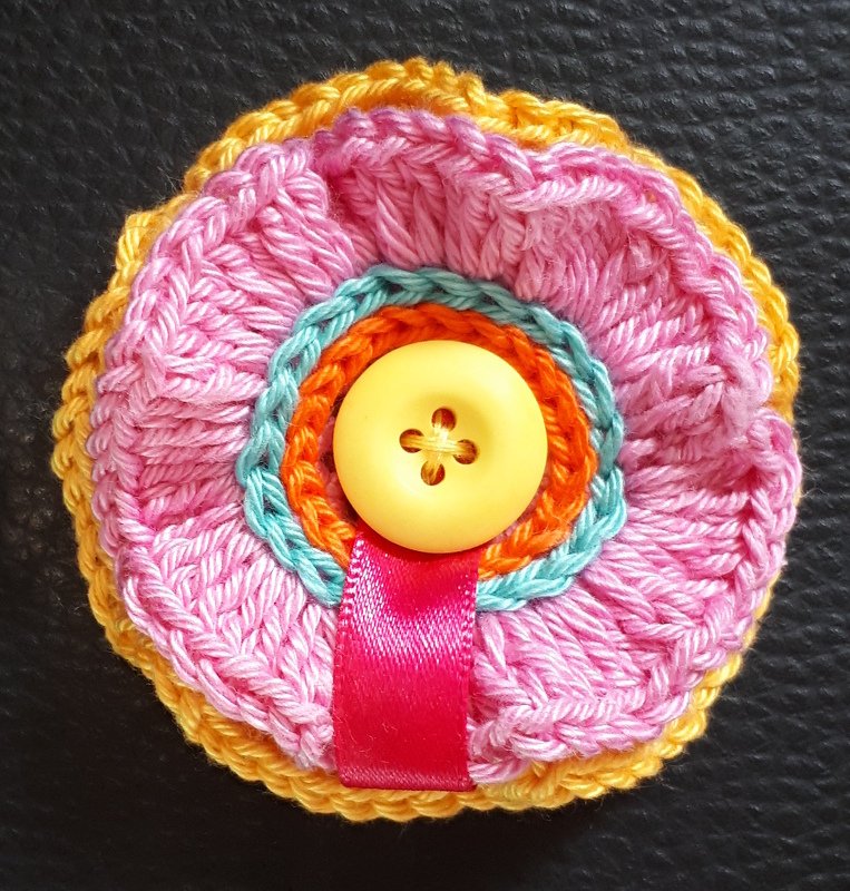 Mehrschichtige gehäkelte Blumenapplikation in Rosa, Türkis, Orange und Gelb mit gelbem Knopf und pinkem Satinband auf dunklem, strukturiertem Hintergrund.