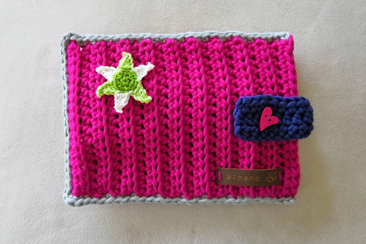 Rechteckige fuchsiafarbene Häkelhülle mit hellgrauem Rand, grün-weißer Stern-Applikation und dunkelblauem Riegel mit pinkem Herzknopf.