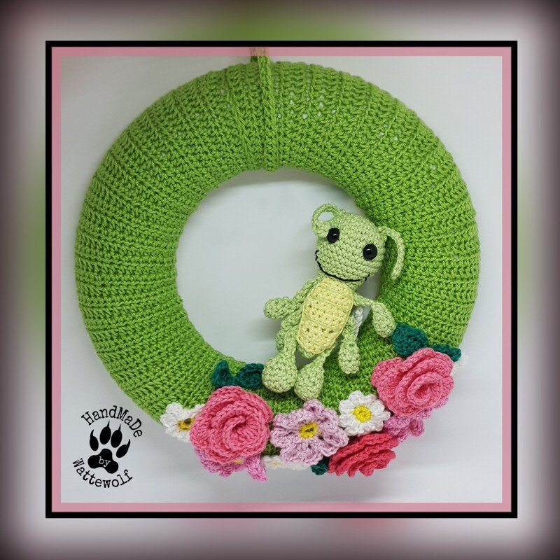 Grüner gehäkelter Türkranz mit kleinem Amigurumi-Frosch und rosa sowie weißen Häkelblumen