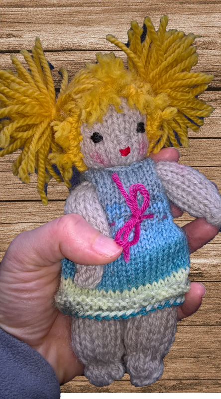 Nahaufnahme einer handgestrickten Amigurumi-Puppe mit gelben Zöpfen, blau-türkisem Kleid und pinker Schleife, gehalten in einer Hand.