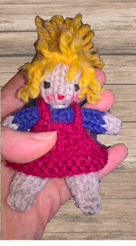Kleine handgestrickte Amigurumi-Puppe mit pinkem Kleid, blauem Oberteil und gelbem Haar in einer Hand vor Holz.