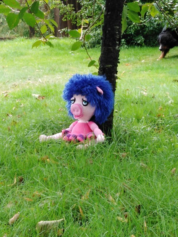 Handgehäkeltes rosa Schwein mit blauen Haaren und lila Rock sitzt im grünen Gras neben einem Baum.