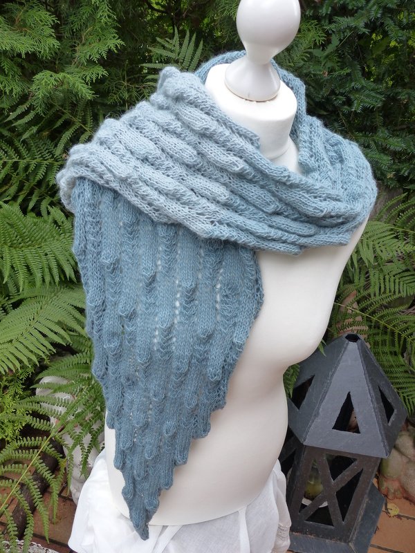 Gestricktes blaugraues Dreieckstuch mit Rippen- und Noppenmuster auf einer Schneiderbüste vor grünen Farnen.