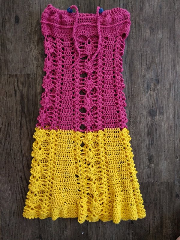 Gehäkeltes zweifarbiges Kinderkleid (Pink oben, Gelb unten) auf dunklem Holzboden liegend