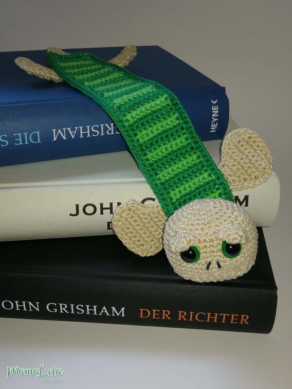 Gehäkeltes grünes Lesezeichen mit beigem Kopf und großen grünen Augen auf einem Bücherstapel.