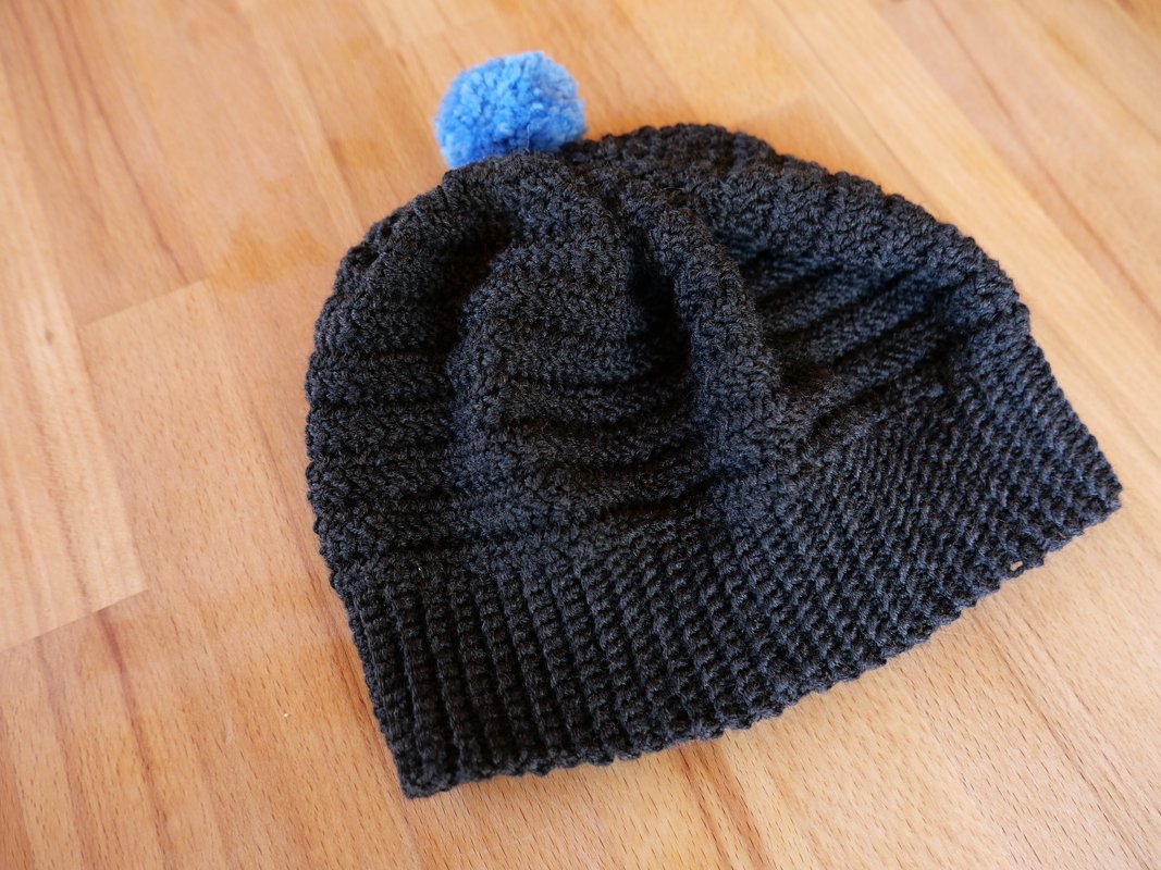 Dunkelgraue gehäkelte Mütze mit blauem Bommel auf Holz