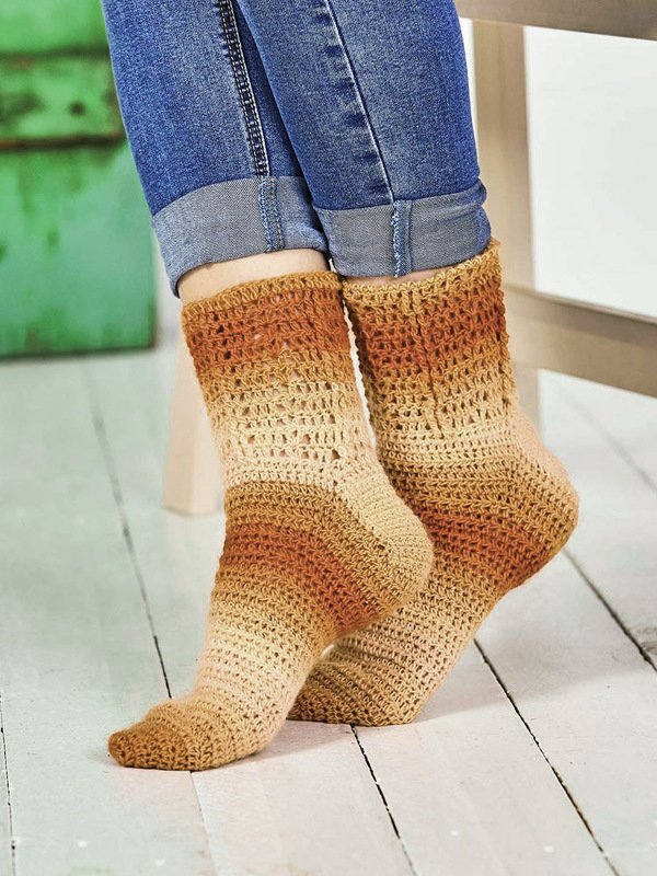 Hochgekrempelte Jeans mit handgehäkelten Socken in Braun‑Beige auf hellem Holzboden