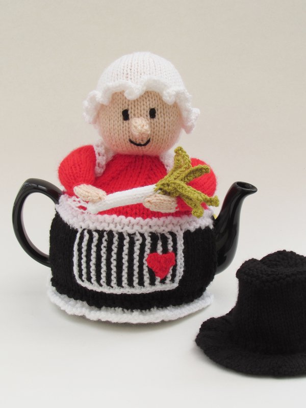 Knitted tea cozy shaped like a character wearing a white bonnet, red top, and black apron, holding a broom, covering a black teapot with a black knitted hat nearby.