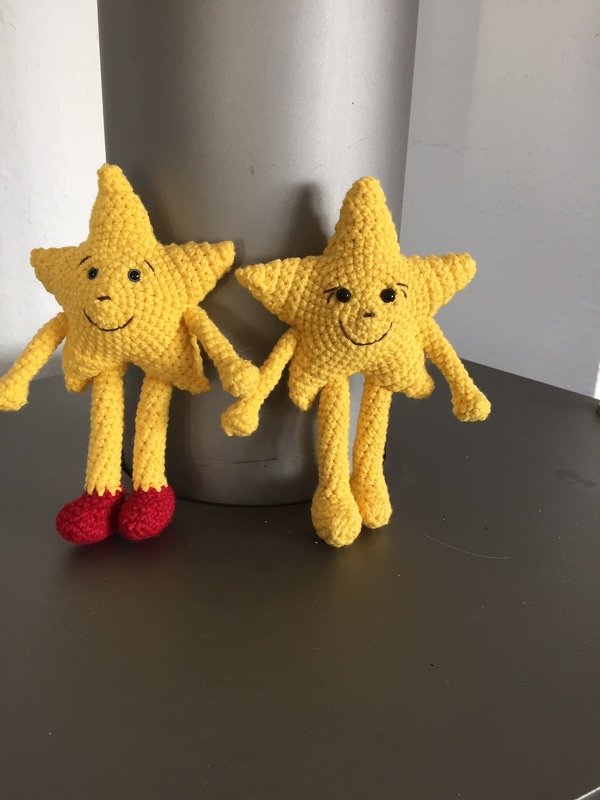 Zwei gelbe, gehäkelte Stern-Amigurumi mit Armen und Beinen und lächelnden Gesichtern, eines mit roten Schuhen