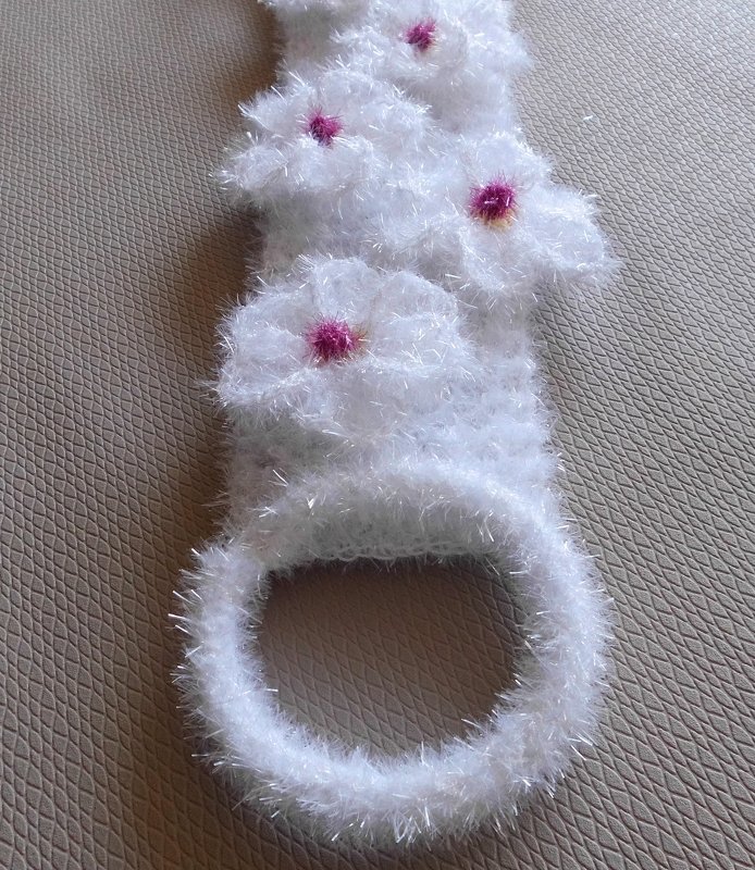 Gehäkelter weißer Handtuchhalter aus flauschiger Wimpernwolle mit fuchsiafarbenen Blüten, liegend auf beigem Untergrund