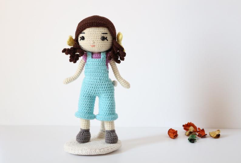 Crocheted amigurumi doll in turquoise overalls with brown pigtails and yellow bows standing on a round base