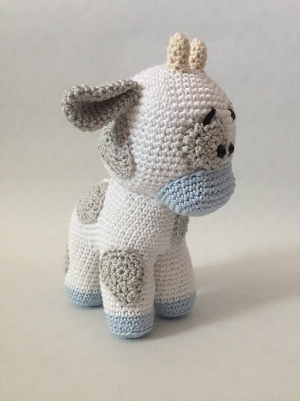 White crocheted amigurumi calf with grey spots and light blue snout and hooves on a neutral background