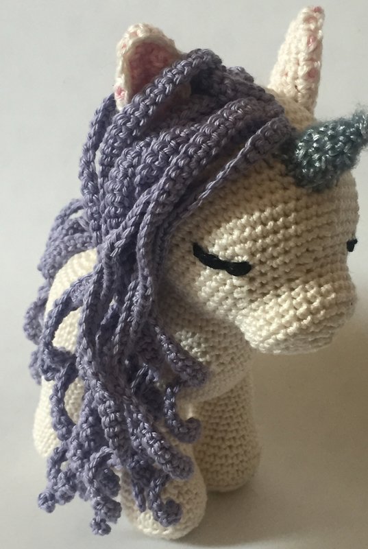 Crocheted amigurumi unicorn toy with cream body, purple mane, grey horn, and pink inner ears.