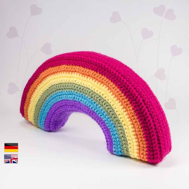 Arched crochet rainbow cushion in layered bright stripes, styled on a pale background with subtle heart motifs