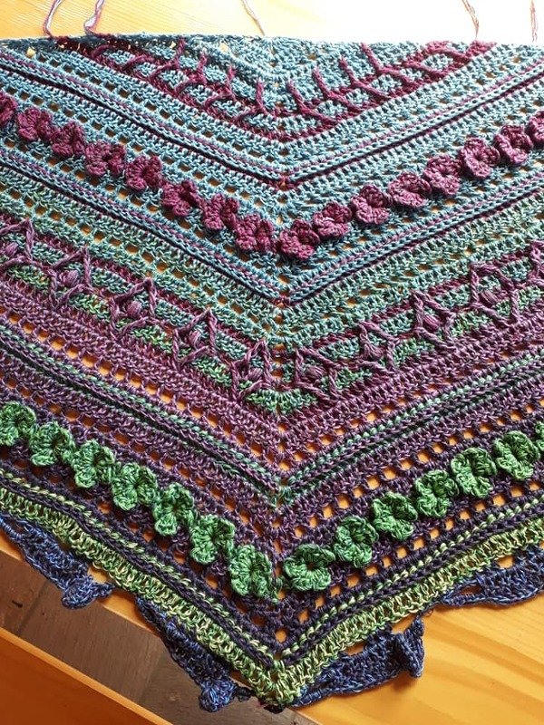 Close-up of a crocheted triangle shawl in blue, green, and purple showing openwork lace, textured stitch rows, and a scalloped edge.