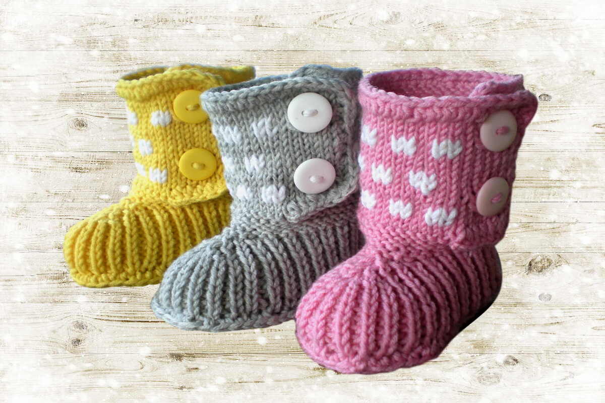 Drei gestrickte Babystiefel in Gelb, Grau und Rosa mit weißen Herzmotiven und seitlichen Knöpfen auf hellem Holzhintergrund.