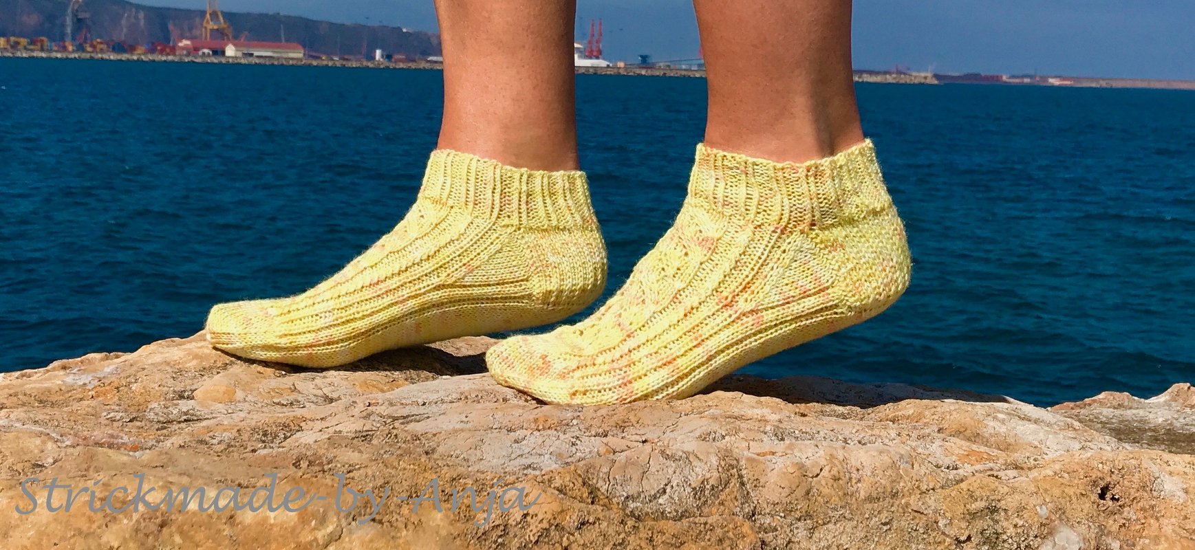 Füße in gelben handgestrickten Knöchelsocken auf Felsen vor blauem Meer
