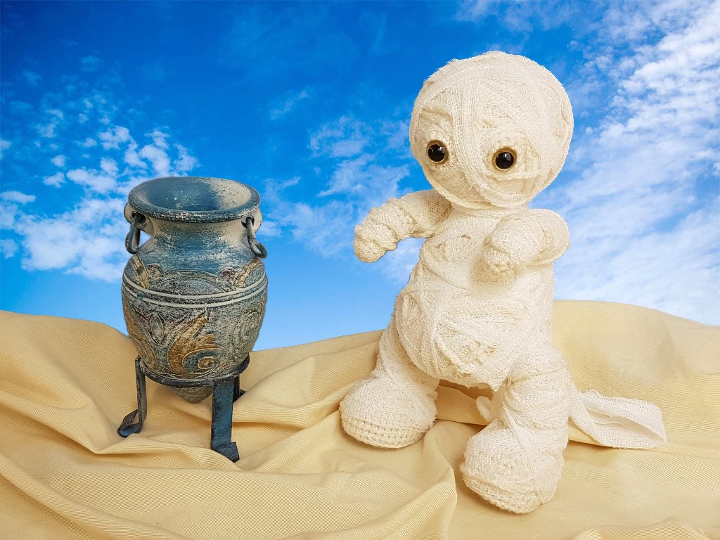 Beige gehäkelte Mumienpuppe mit dunklen Augen neben verzierter Vase vor blauem Himmel