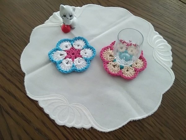 Zwei gehäkelte Blumen-Untersetzer in Blau
‑Weiß und Rosa
‑Beige auf runder bestickter Tischdecke mit Glas und kleiner Katzenfigur